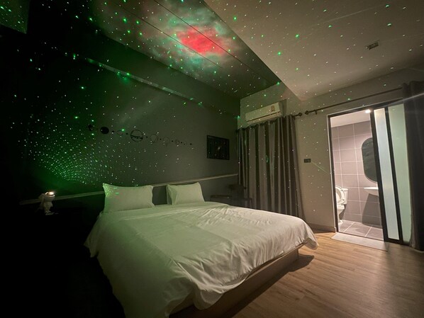 Un dormitorio con cama, pared con diseño de galaxia y baño visible a través de una puerta de cristal.