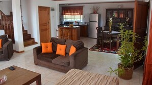 A cozy living room with a brown sofa, a wooden coffee table, and a dining area with a table and chairs. A kitchen with a refrigerator and a sink is visible in the background.