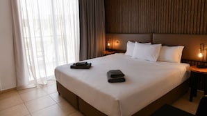 A hotel room with a large bed, two bedside tables with lamps, a window with sheer curtains, and a wooden panel wall. - King's Holiday Apartments (Paphos)