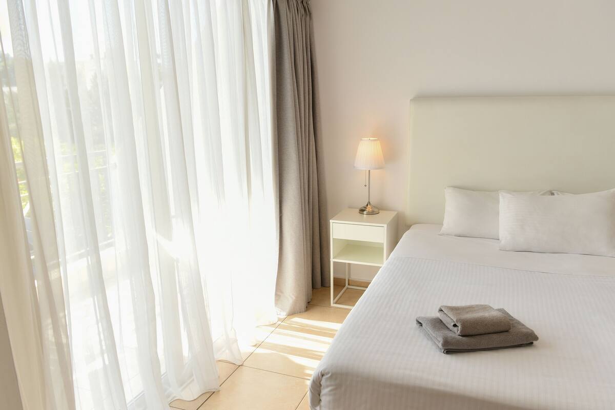 A hotel room with a bed, a nightstand with a lamp, and sheer curtains.