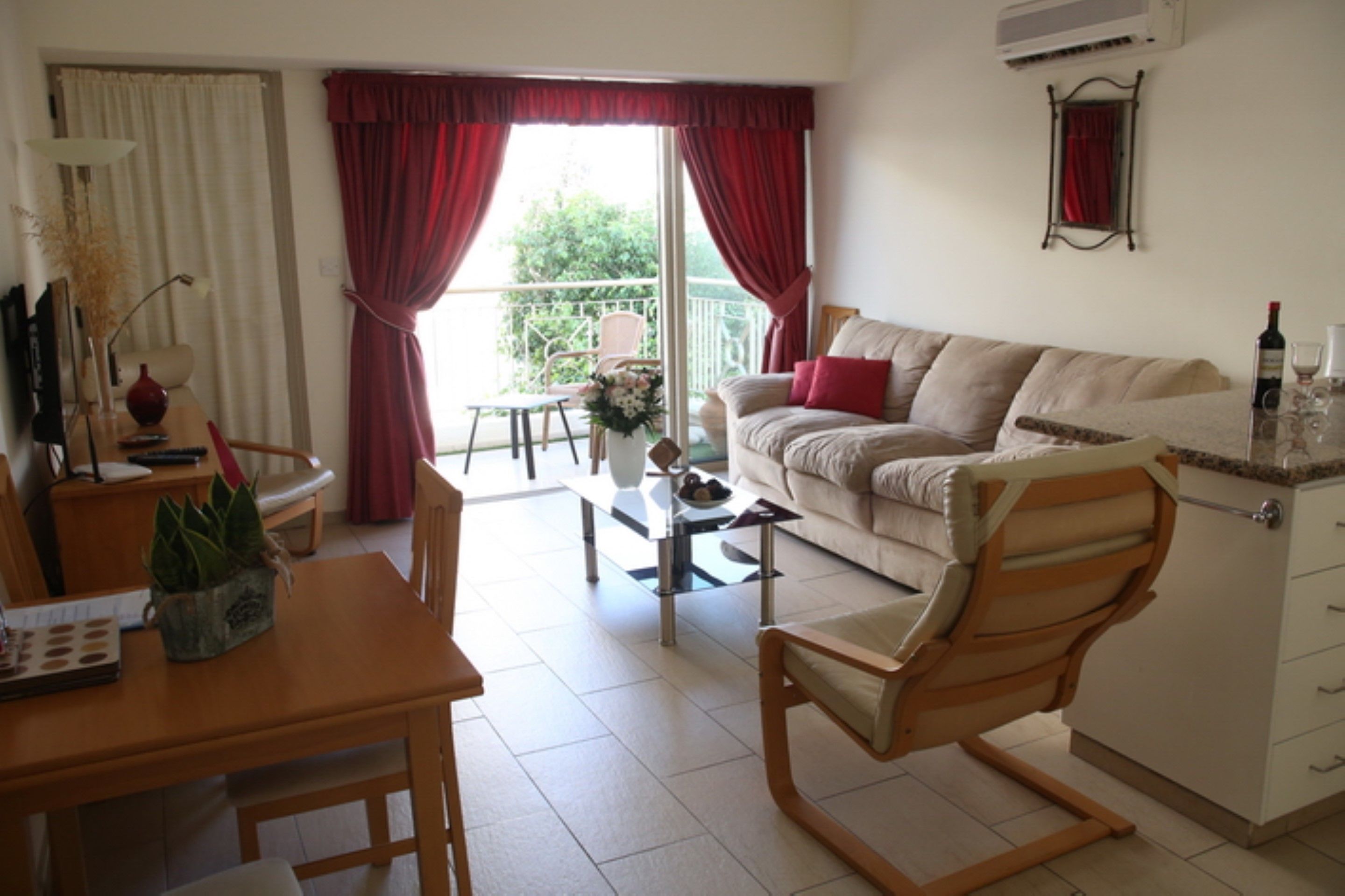 A living room with a sofa, a dining table, and a chair. There is a window with curtains, a plant on the table, and a vase.