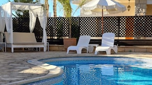 A swimming pool with a decorative edge, white lounge chairs, and a pergola with white fabric. - King's Holiday Apartments (Paphos)