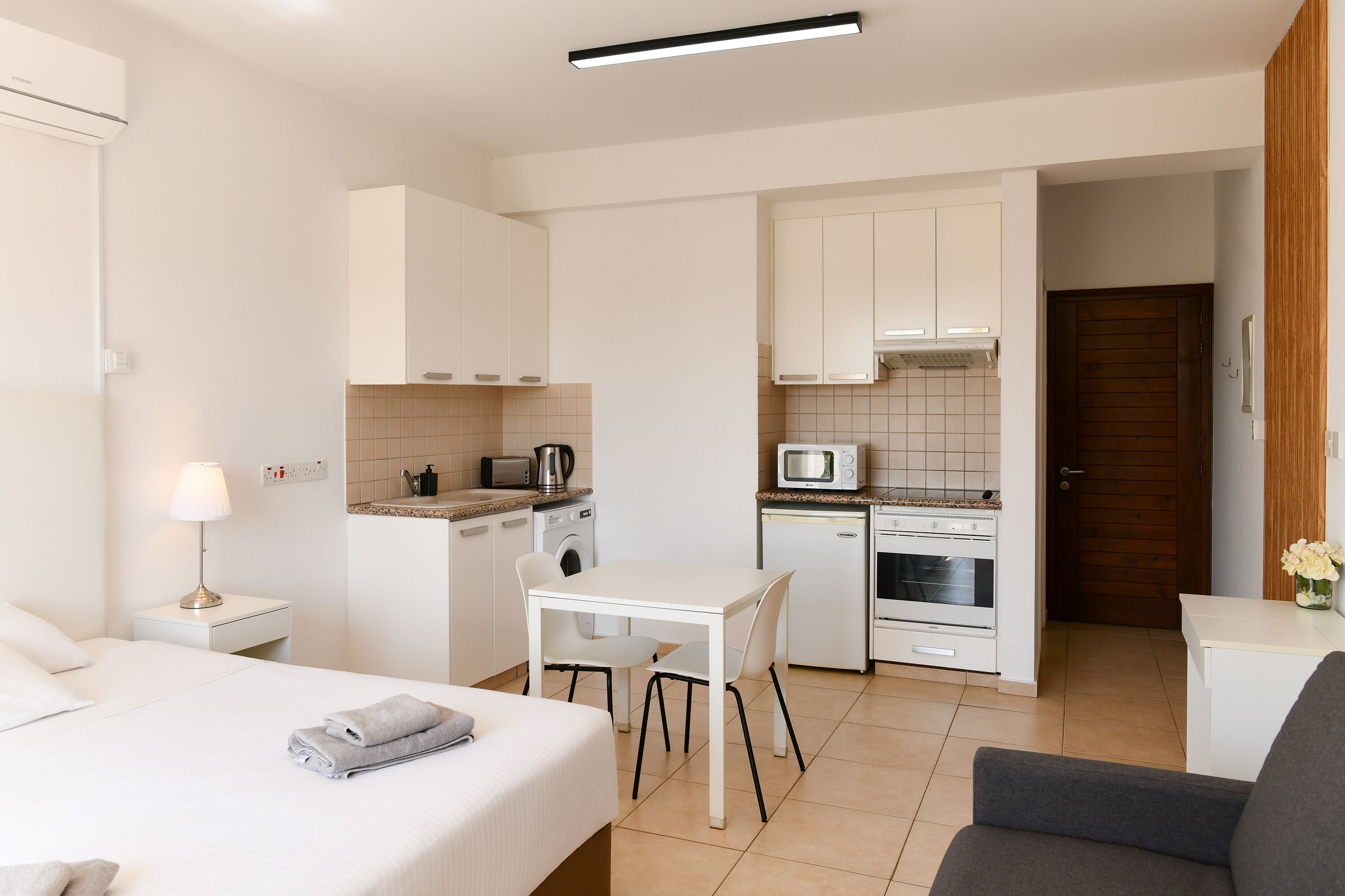 A compact hotel room with a bed, a small table, a chair, a kitchenette, and a bathroom.