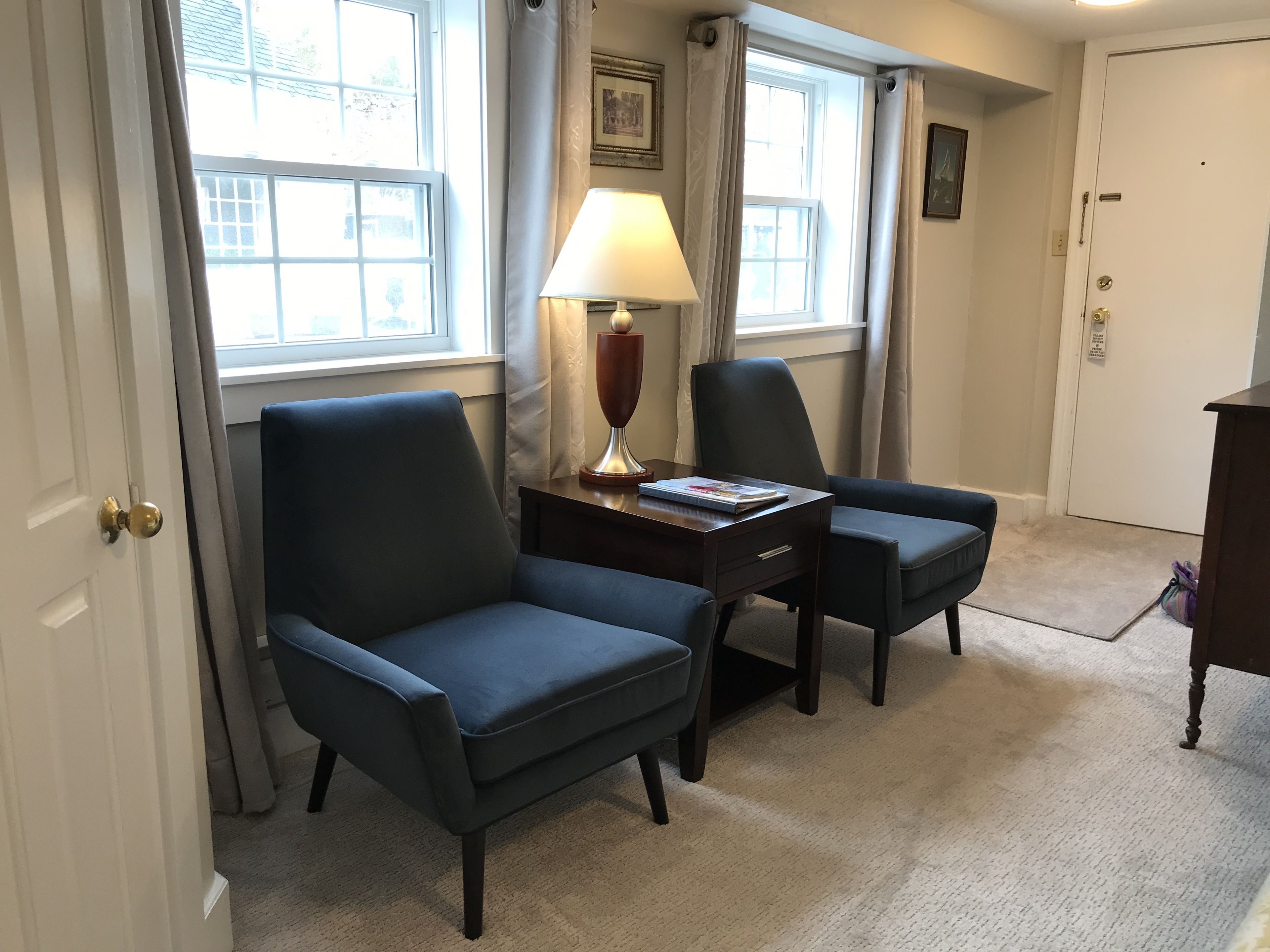 A room with two dark blue armchairs, a small wooden table with a lamp, and a framed picture on the wall.
