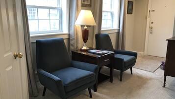 A room with two dark blue armchairs, a small wooden table with a lamp, and a framed picture on the wall.