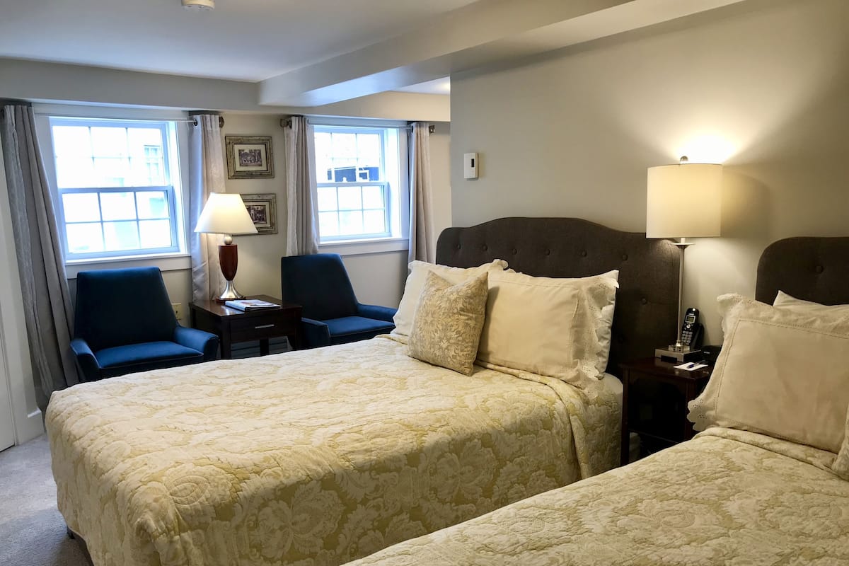 A hotel room with two beds, a desk, a chair, a lamp, and a window with curtains.