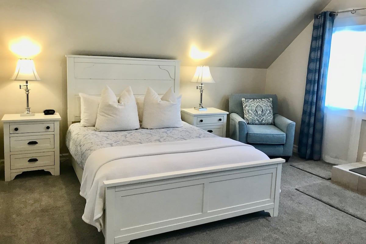 A bedroom with a white bed, two bedside tables with lamps, a chair with a patterned cushion, and a window with curtains.