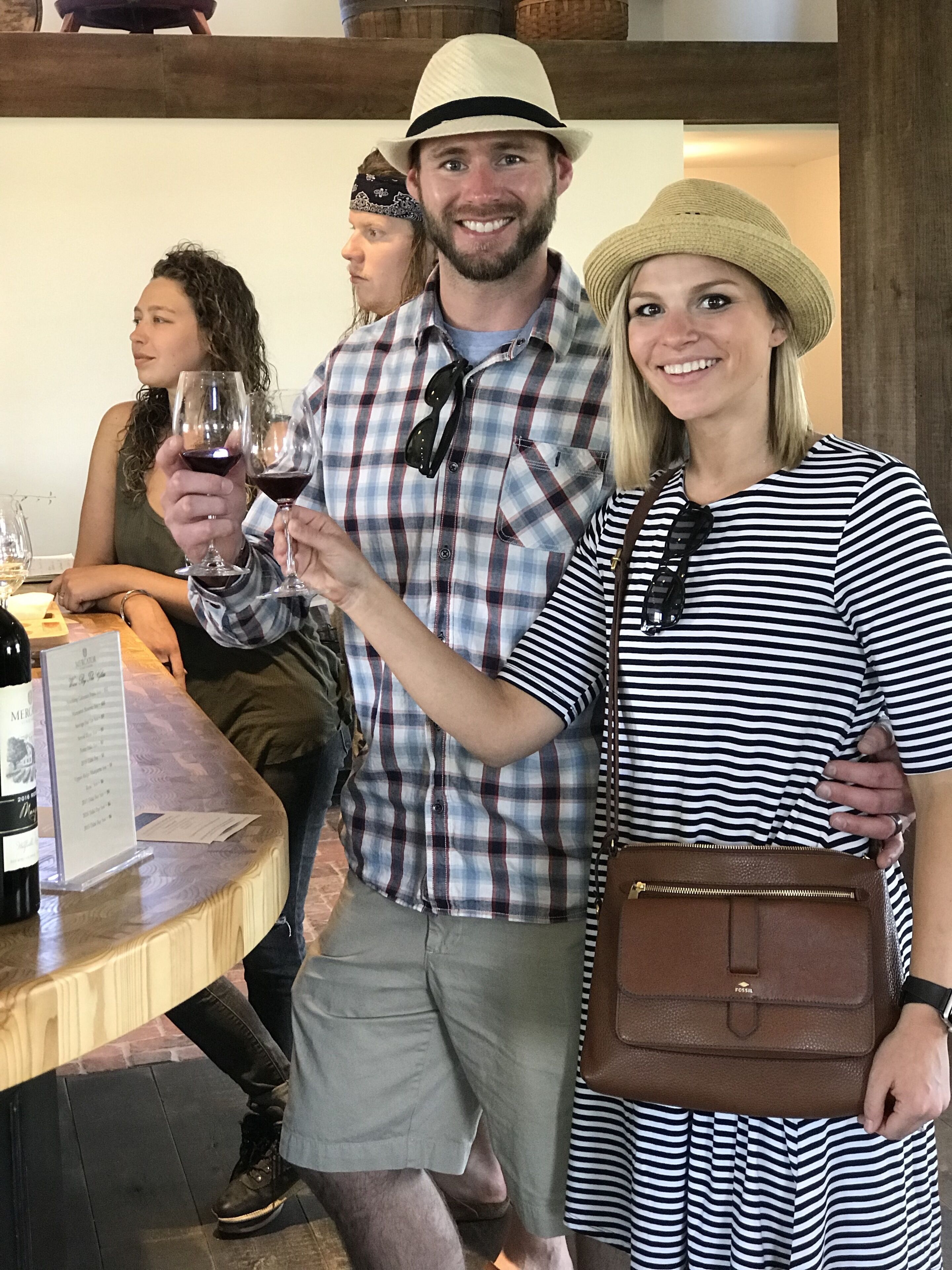 A group of people at a wine tasting event, with a wooden table displaying wine bottles and glasses.
