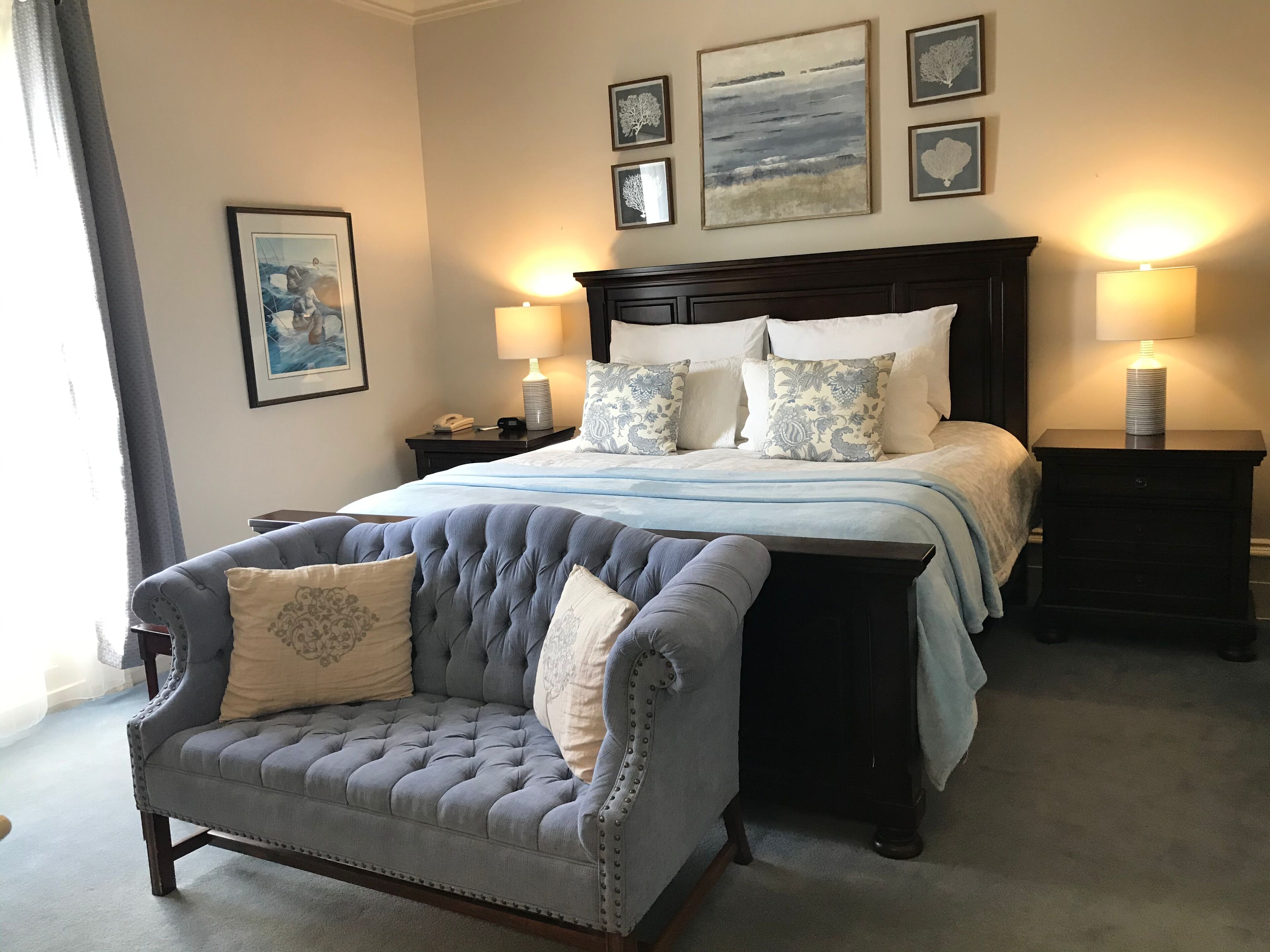 A neatly arranged bedroom with a large bed, two bedside tables with lamps, a tufted armchair, and a framed picture on the wall.