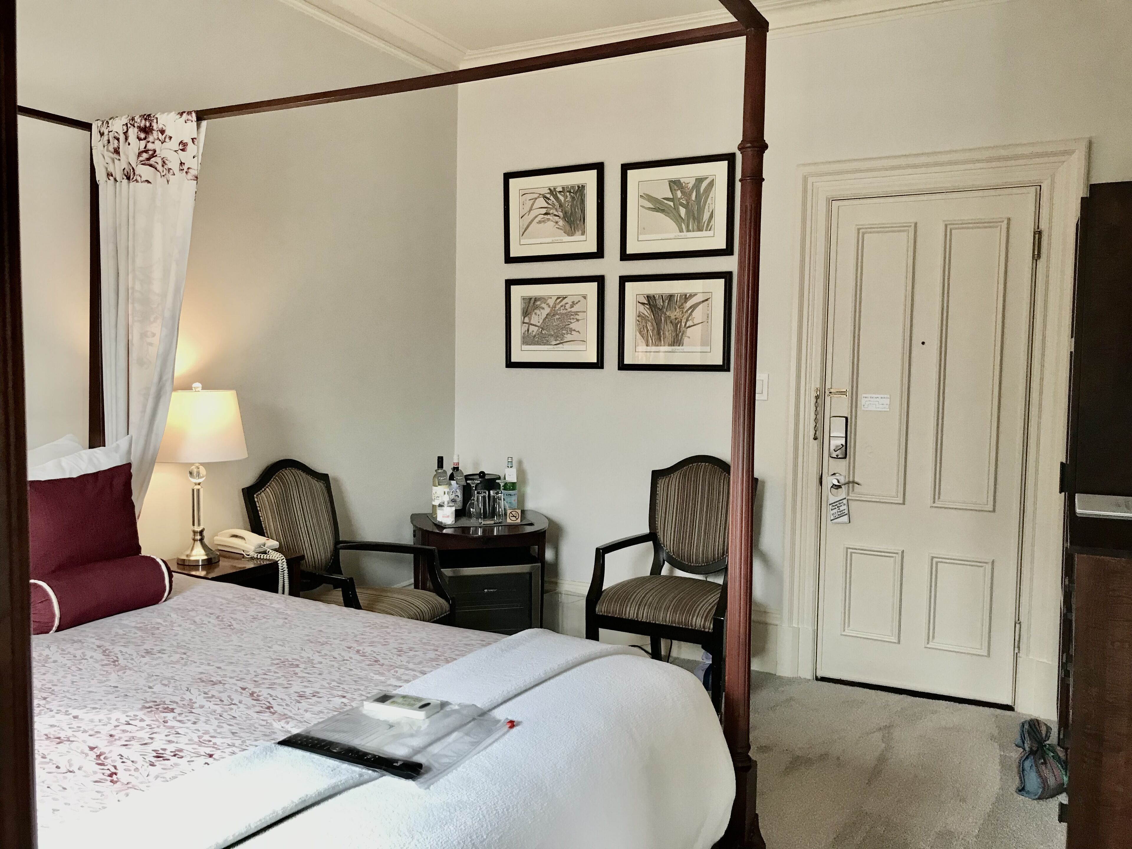 Un lit à baldaquin avec une couvre-lit blanche, une table de chevet avec une lampe, une chaise et un bureau. Le mur est décoré d’illustrations botaniques encadrées.