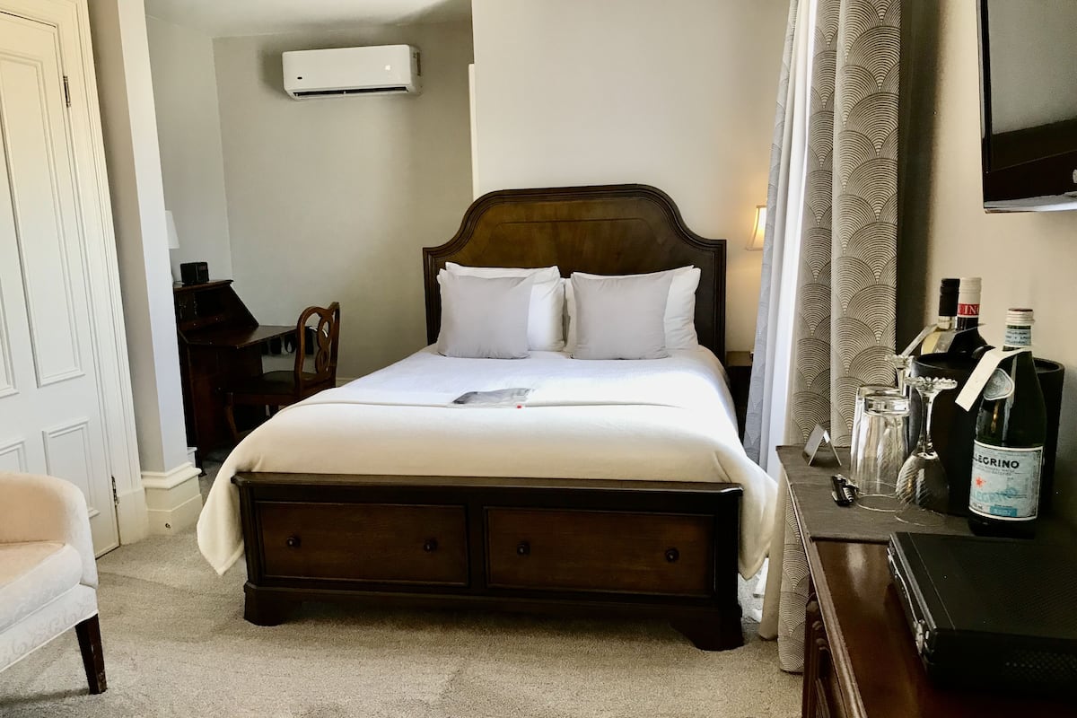 A neatly made bed with white linens, a wooden headboard, and a bedside table with a bottle of wine and glasses.