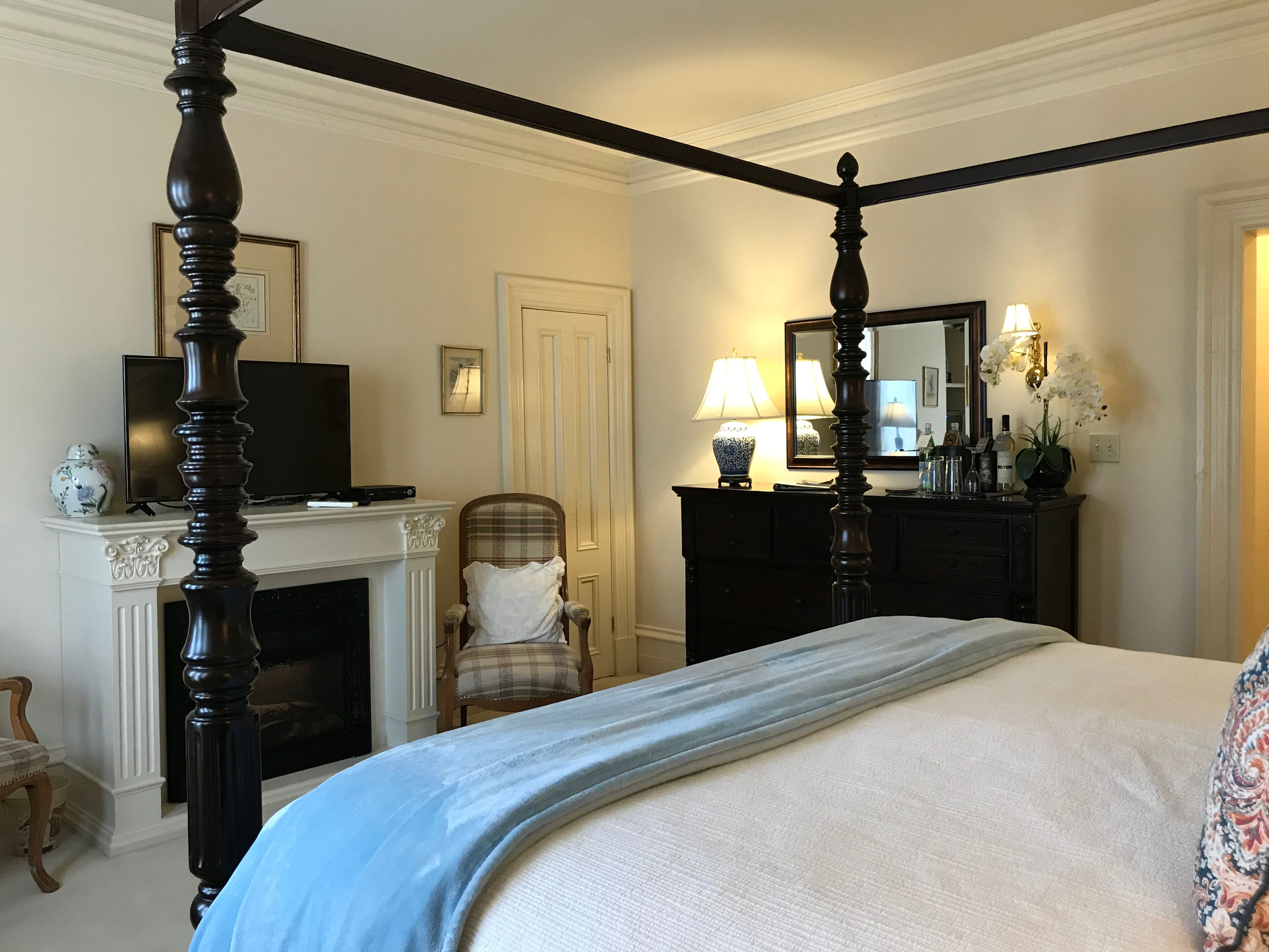 A four-poster bed with a white and blue bedspread, a television on a mantelpiece, a fireplace, a chair, a dresser with a mirror, and a lamp.