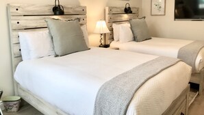 Habitación de hotel con dos camas, cada una con cabecero de madera, ropa de cama blanca y una manta gris. Hay lámparas de pared y cuadros enmarcados sobre las camas.
