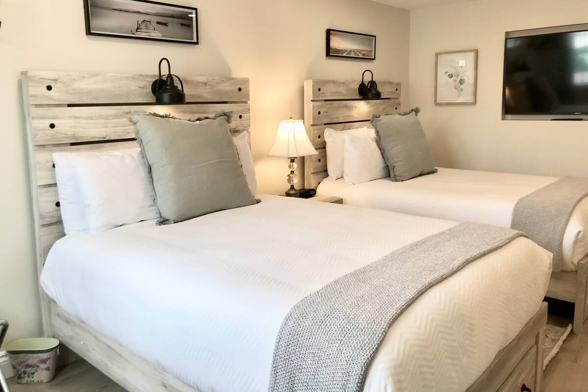 A hotel room with two beds, each with a wooden headboard, white bedding, and a grey throw blanket. There are wall-mounted lamps and framed pictures above the beds.