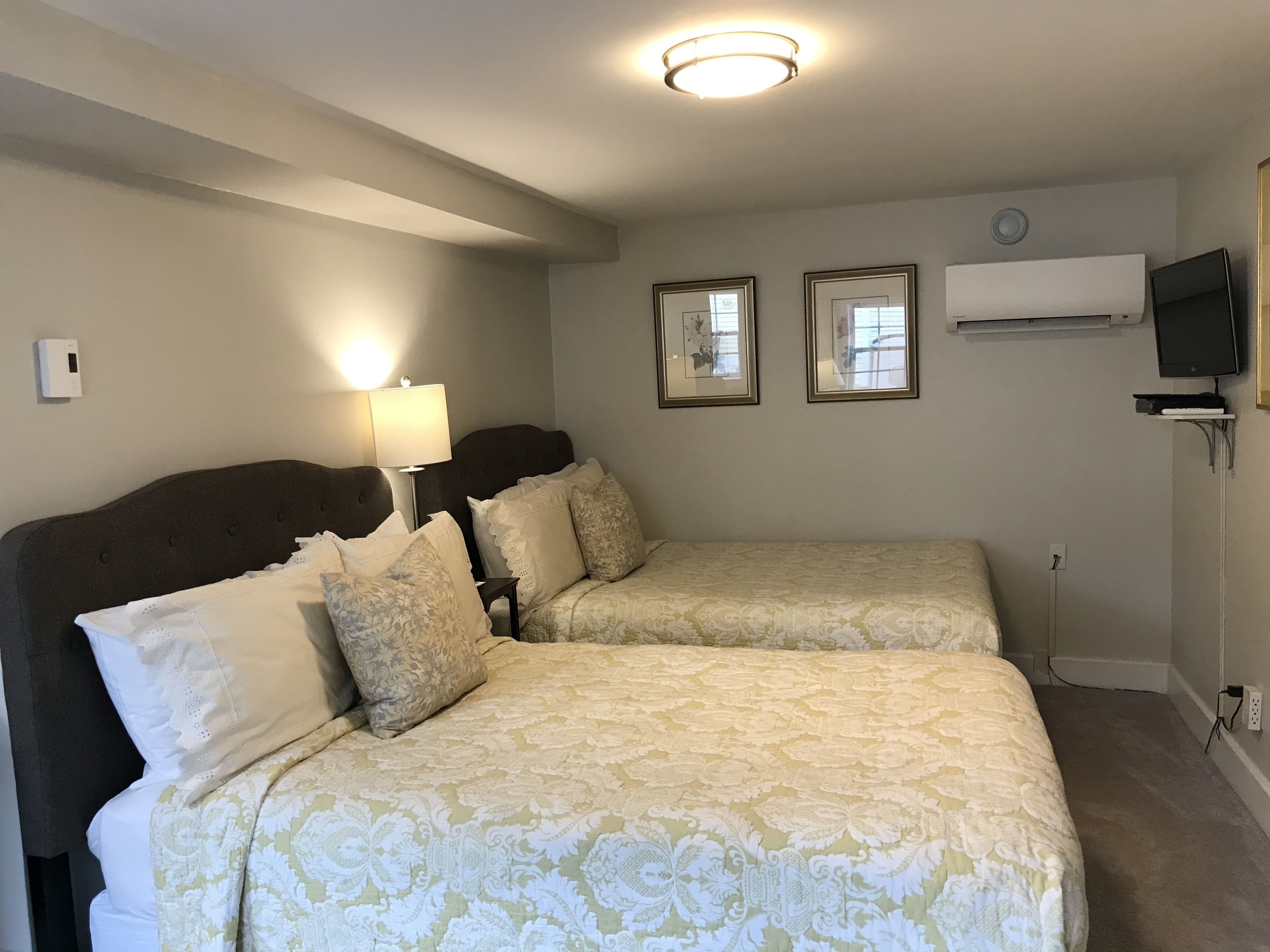 A hotel room with two beds, a bedside lamp, framed pictures on the wall, and a TV mounted on the wall.