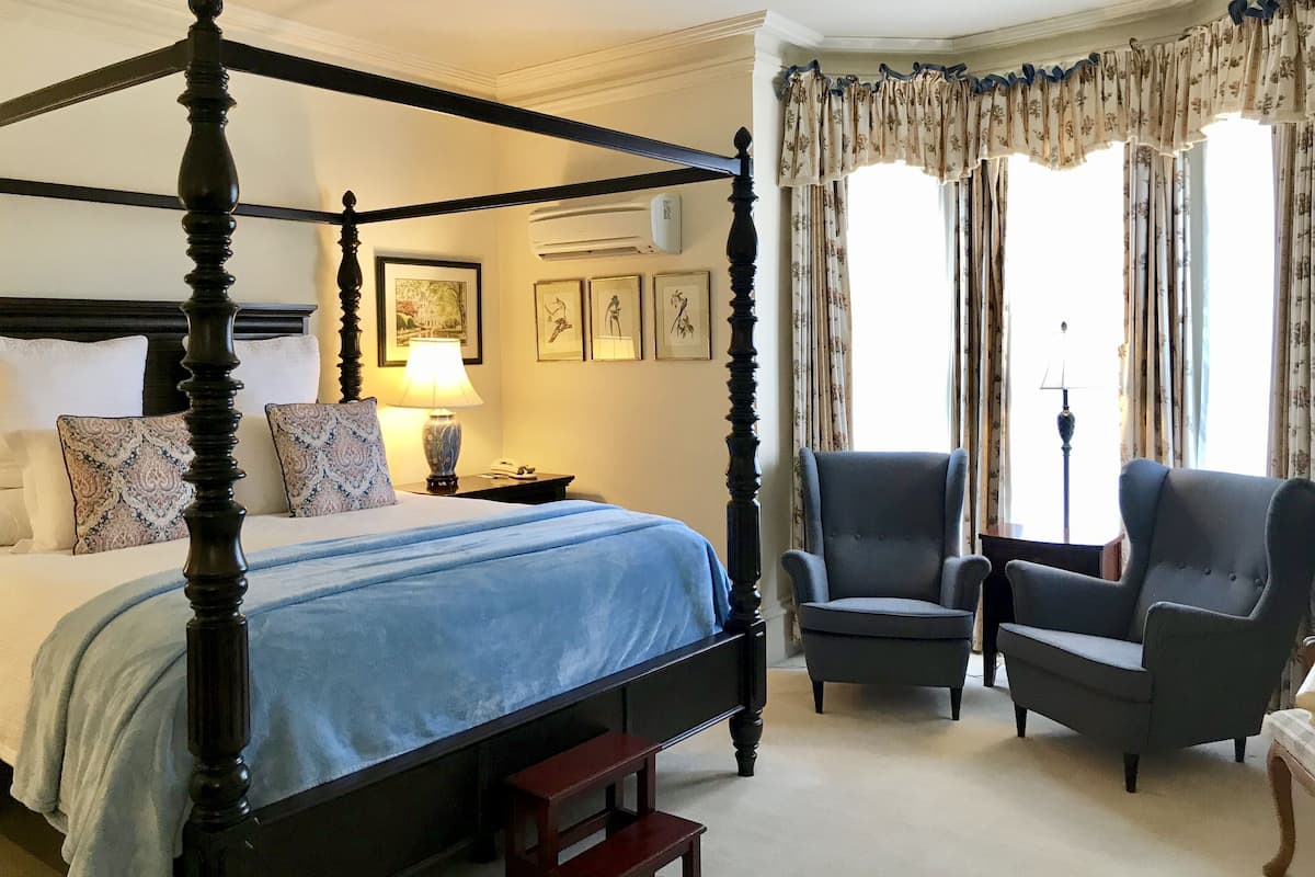 A four-poster bed with a blue bedspread, two armchairs, a nightstand with a lamp, and framed artwork on the wall.