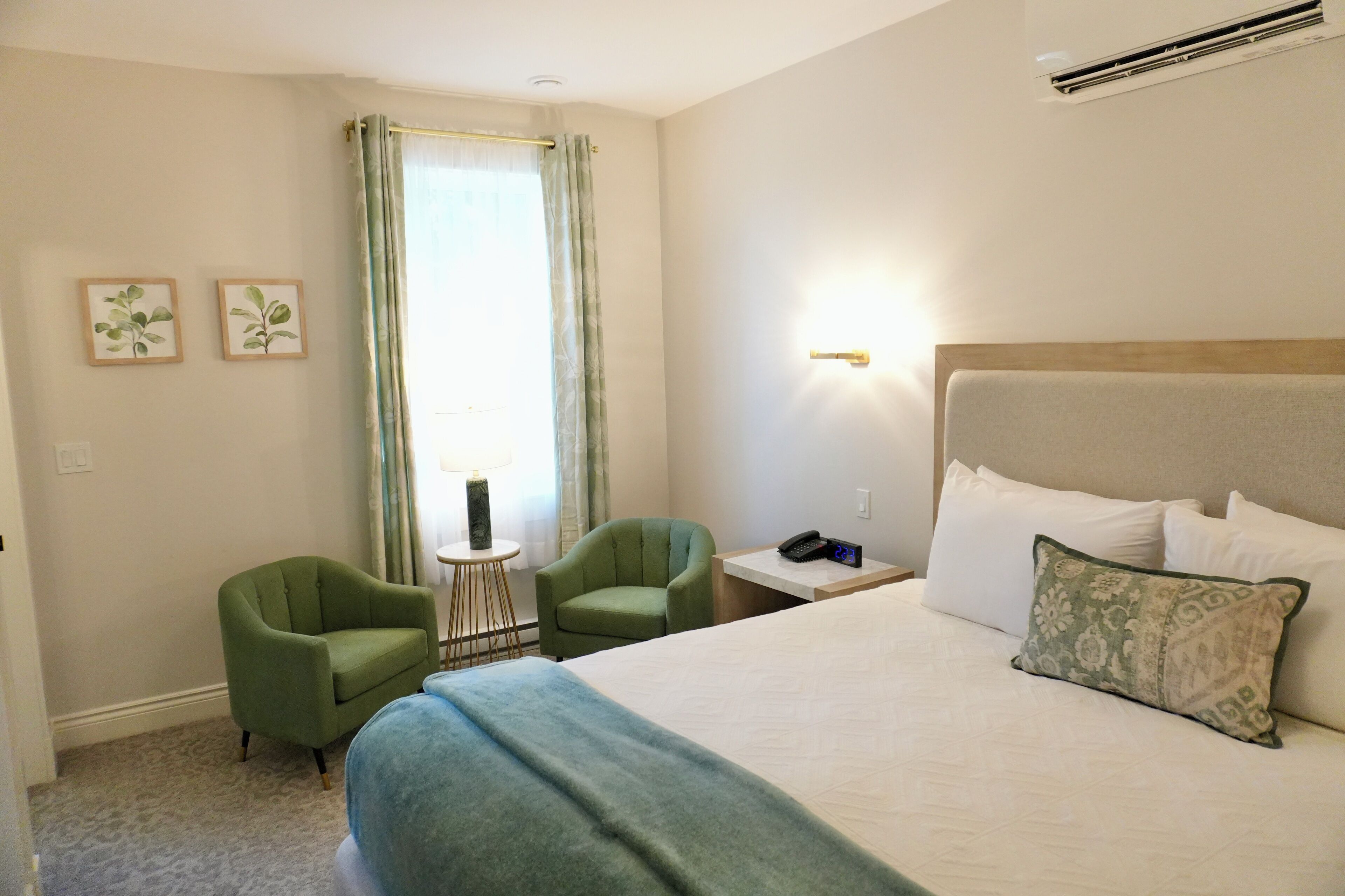 A hotel room with a large bed, two green armchairs, a small table, and a window with curtains.
