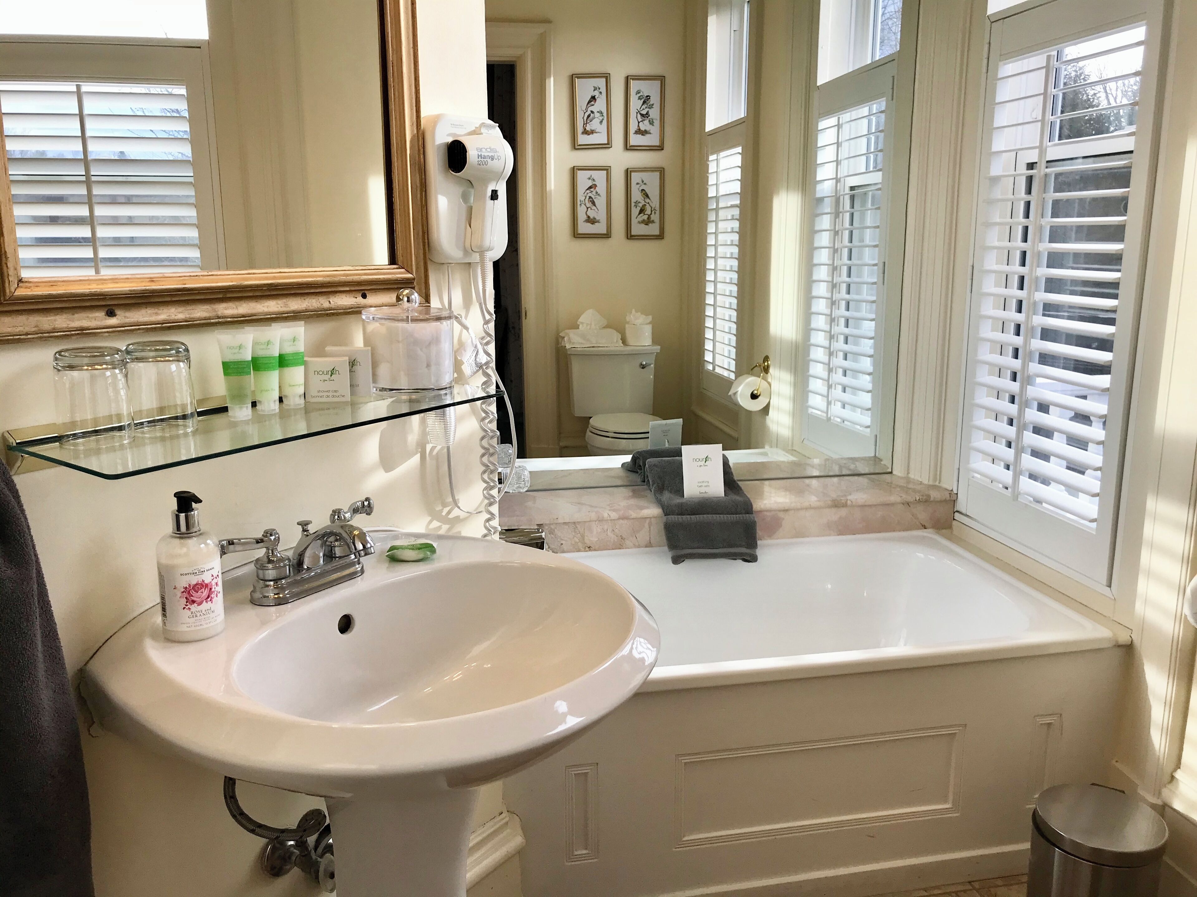 A bathroom with a white sink, a glass shelf with toiletries, a hairdryer, and a window with blinds.