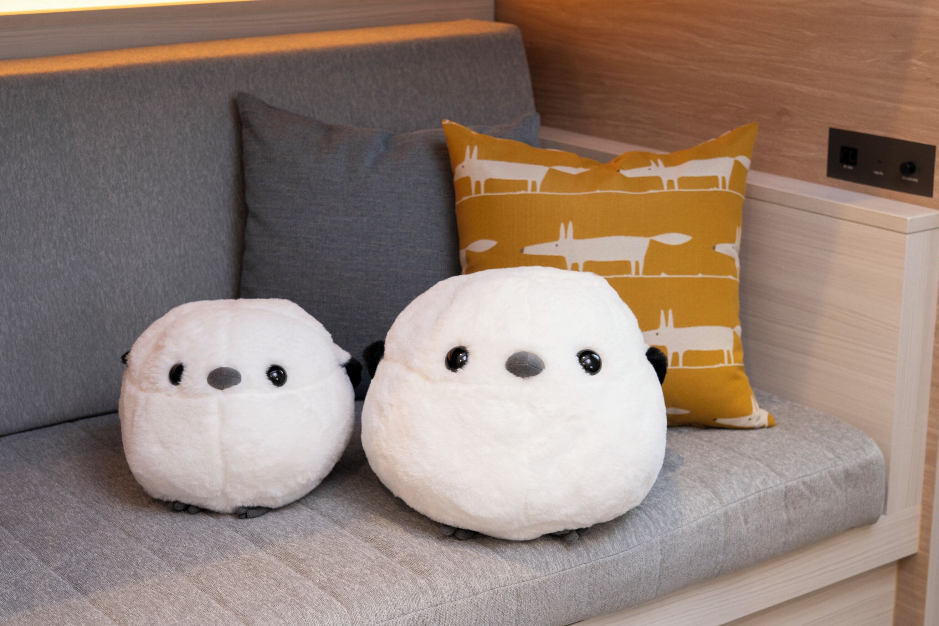 Um sofá cinzento com dois brinquedos de pelúcia brancos e uma almofada amarela mostarda com padrão.