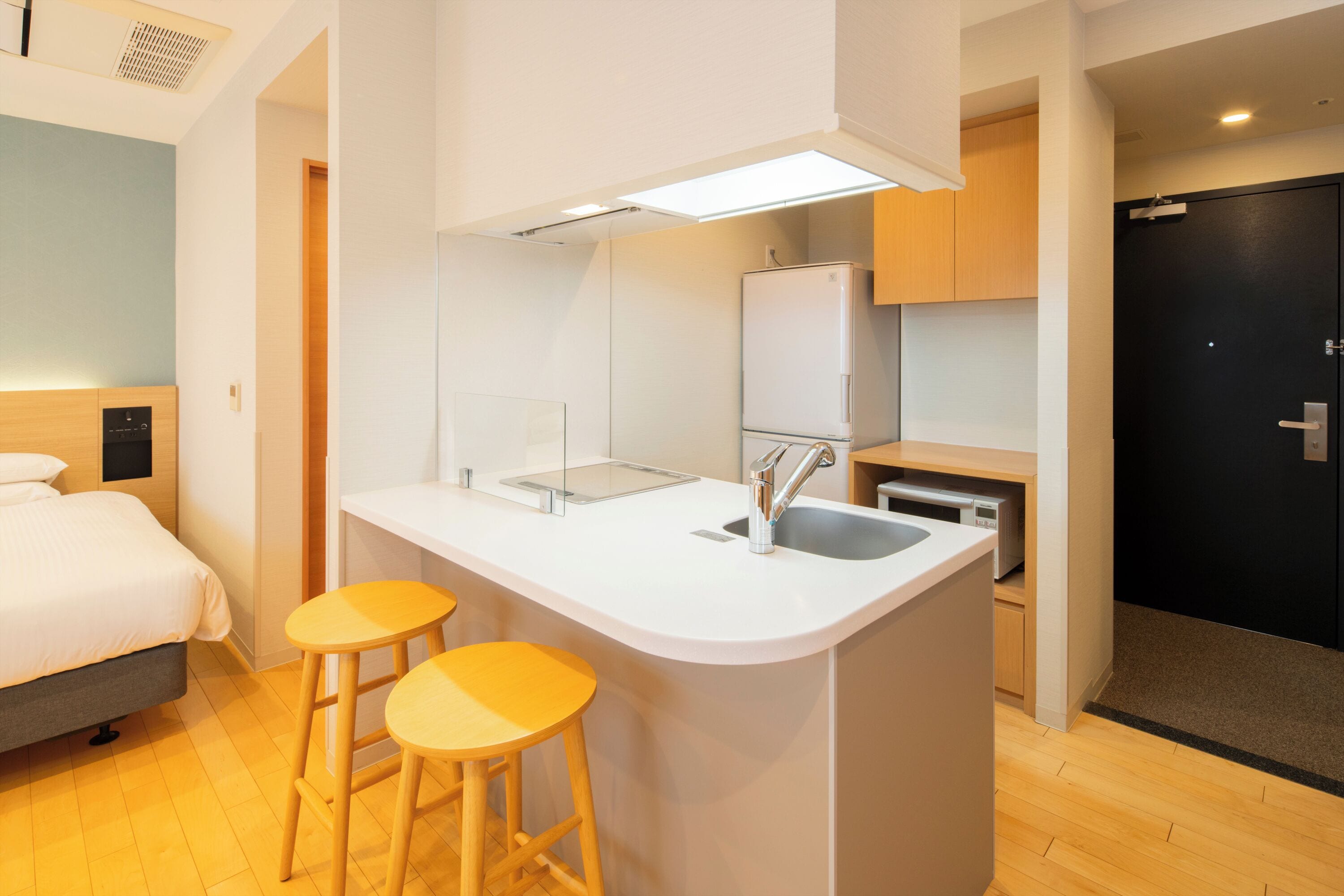 A compact hotel room with a kitchenette, a bed, and two yellow stools.