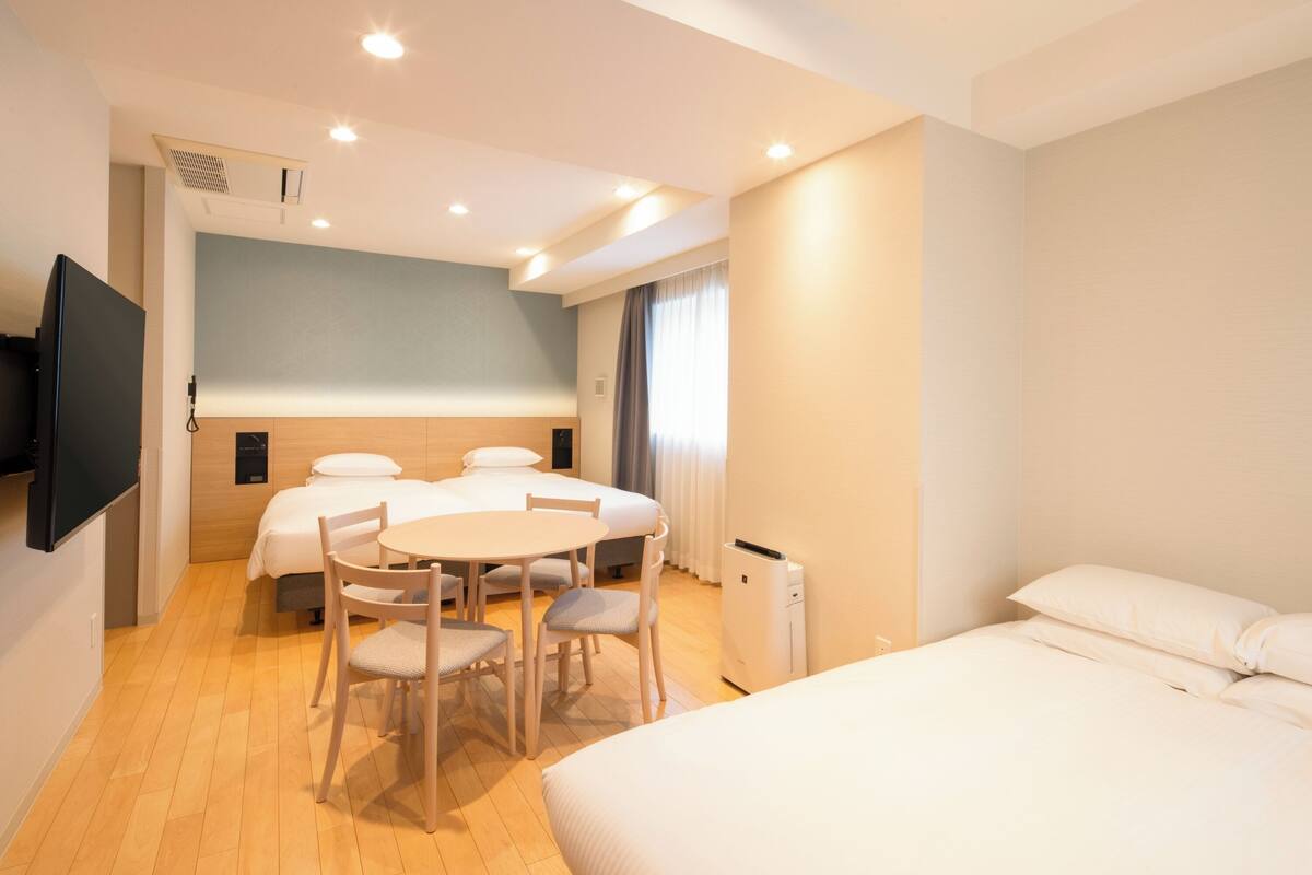 Une chambre d’hôtel comprenant un lit, une table à manger avec des chaises, un téléviseur fixé au mur et une fenêtre avec des rideaux.
