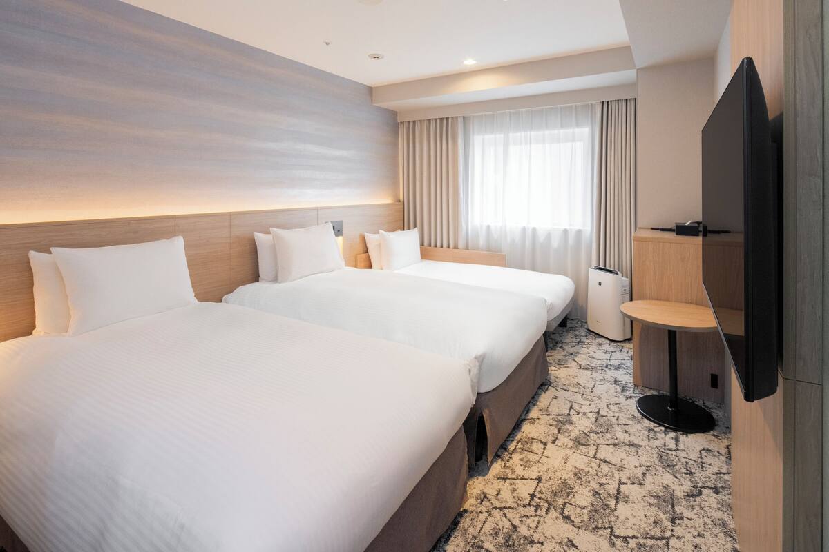 Une chambre d’hôtel avec deux lits, une télévision, une petite table et une grande fenêtre avec des rideaux.