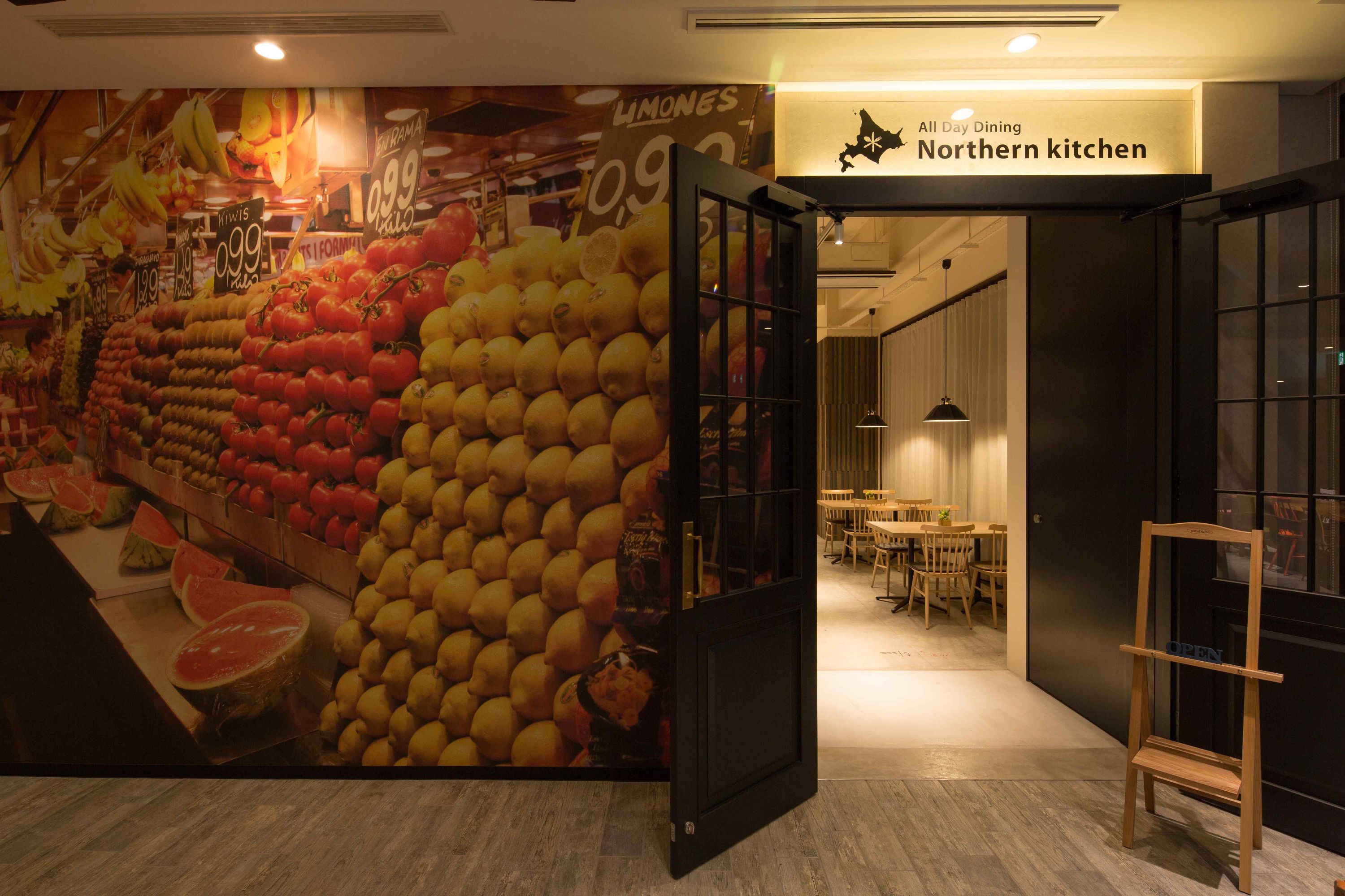 A restaurant interior with a wall mural of fruits and vegetables, a sign for 'All Day Dining Northern kitchen', and a wooden chair in the foreground.
