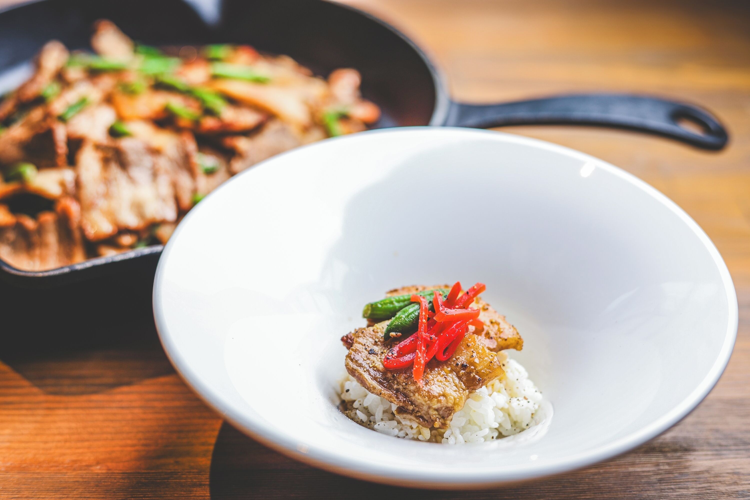 Un bol blanc contenant un morceau de viande et du riz, et une poêle avec des légumes sautés en arrière-plan.
