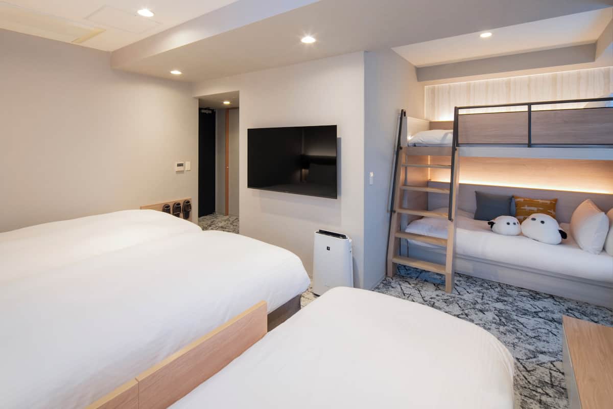 Une chambre d’hôtel avec deux lits, un téléviseur fixé au mur, un lit superposé avec des oreillers et une poubelle.