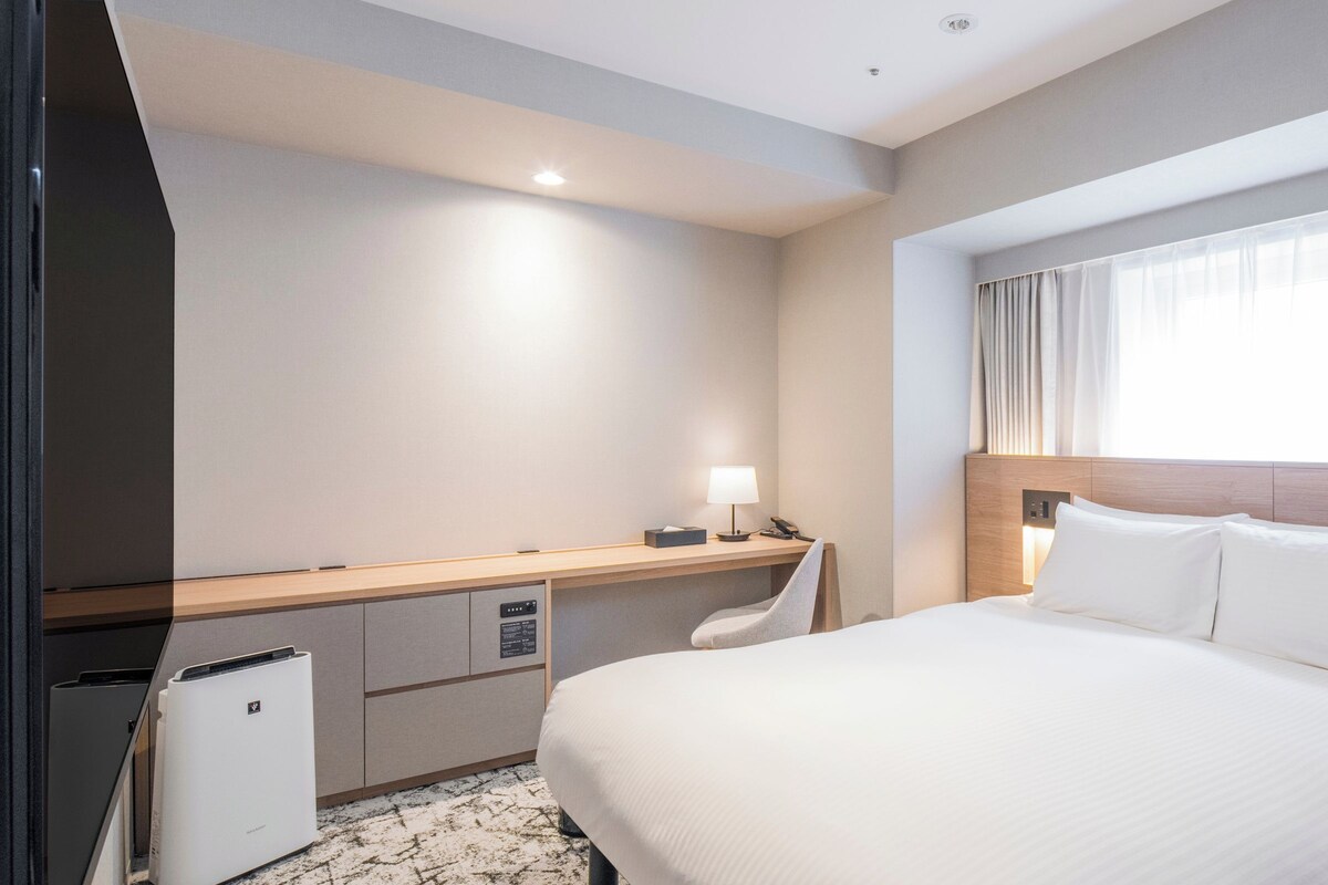A hotel room with a bed, bedside table, lamp, and a small desk with a chair.
