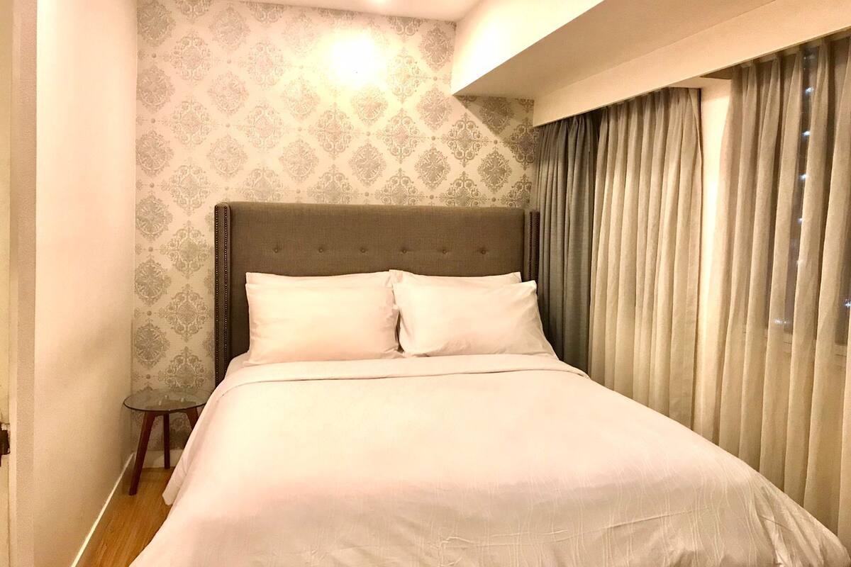 A neatly made bed with a headboard, a small bedside table, and a window with curtains.