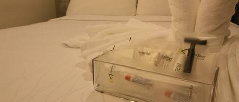 Un récipient transparent en verre contenant des articles de toilette, posé sur une table de chevet recouverte de linge de lit blanc.