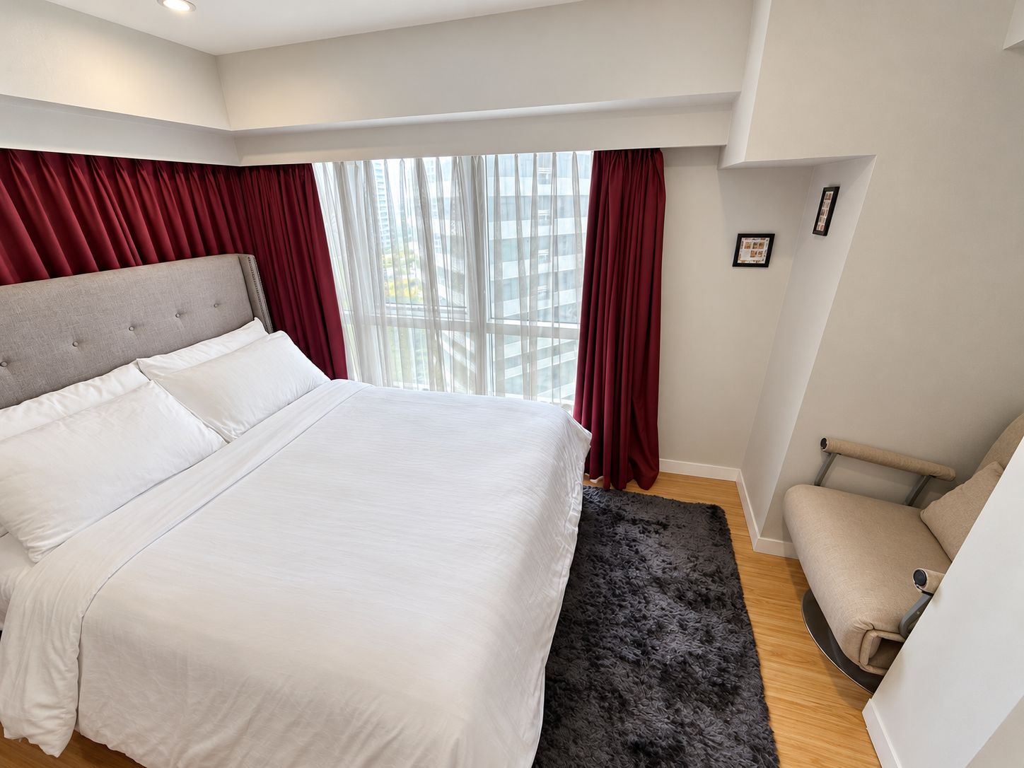 A bedroom with a large bed, a chair, a window with curtains, and a small rug.