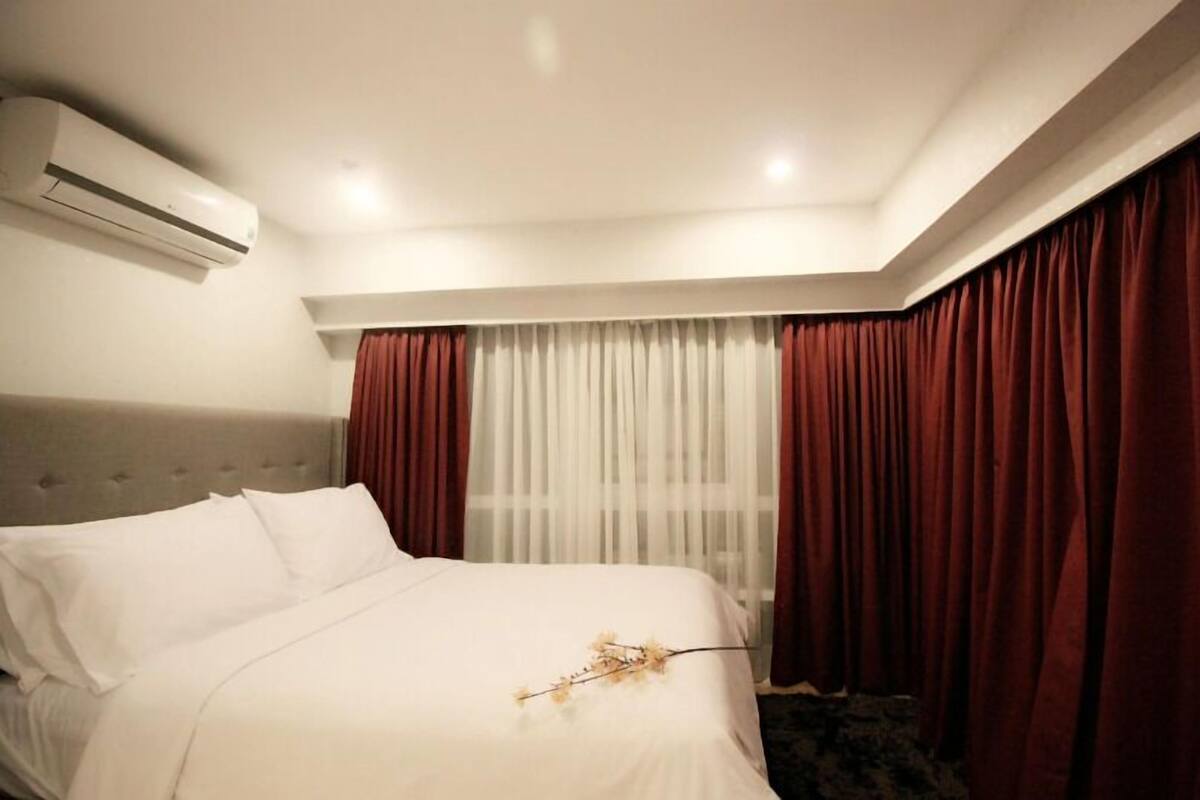 A hotel room with a bed, an air conditioning unit, a window with sheer curtains, and a red curtain.