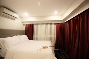 Hotellihuone, jossa on sänky, ikkuna verhoilla, ilmastointilaite ja matolla peitetty lattia.