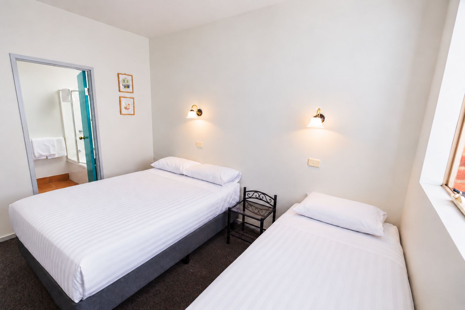 A hotel room with two single beds, a small chair, a mirror, and two wall-mounted lights.