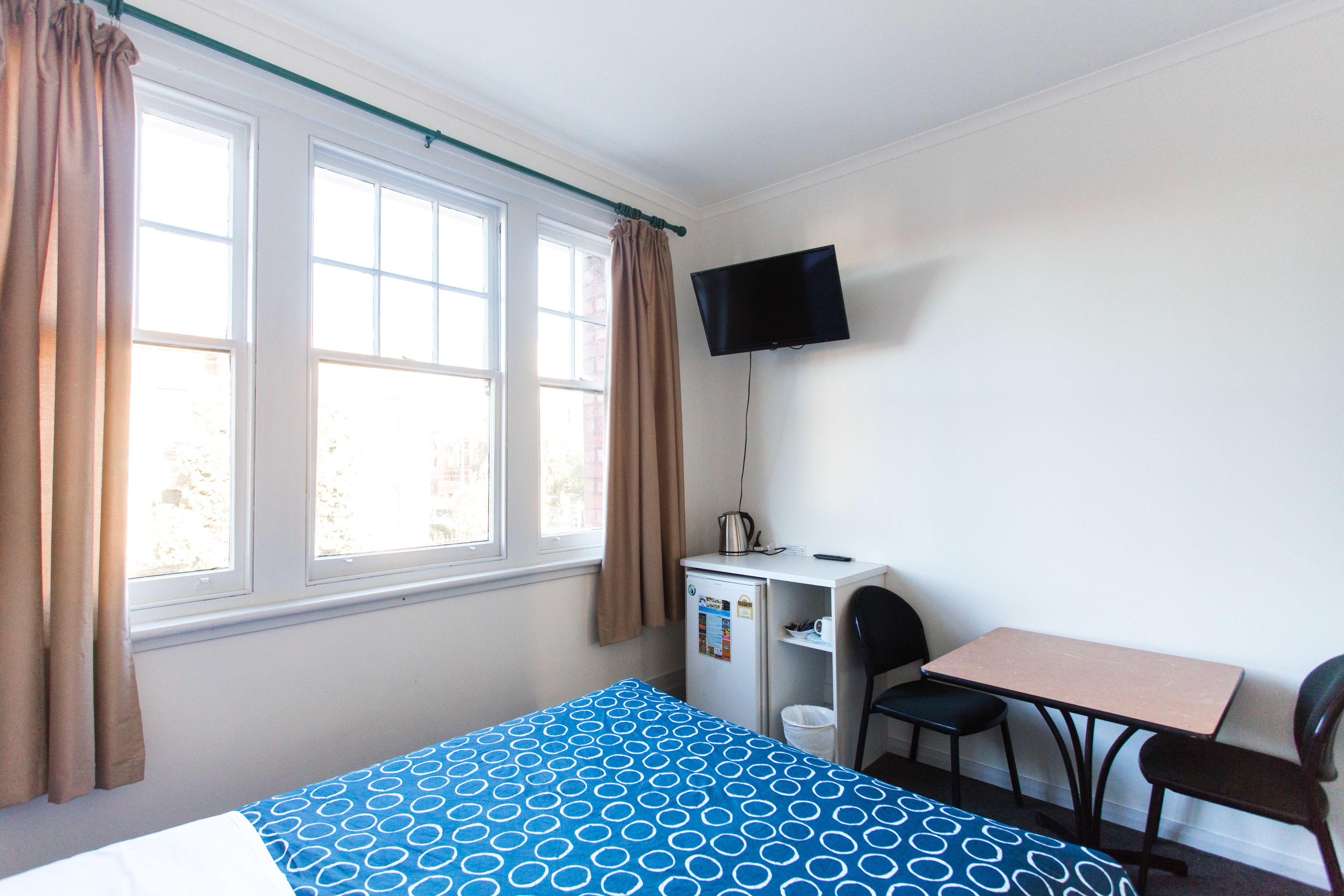 Quarto com cama, escrivaninha, cadeira, televisão e janela com cortinas.