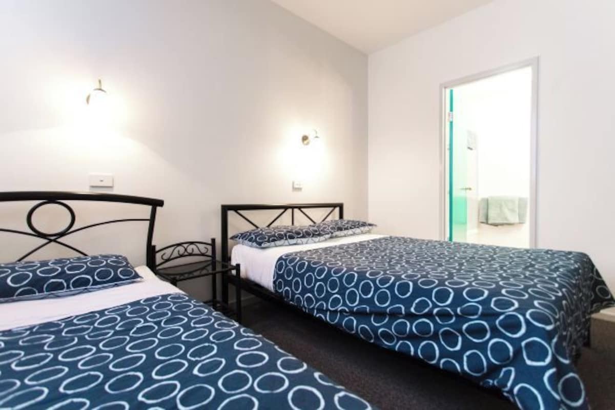 A double room with two beds, each with a patterned duvet cover, a black metal headboard, and a wall-mounted light fixture.