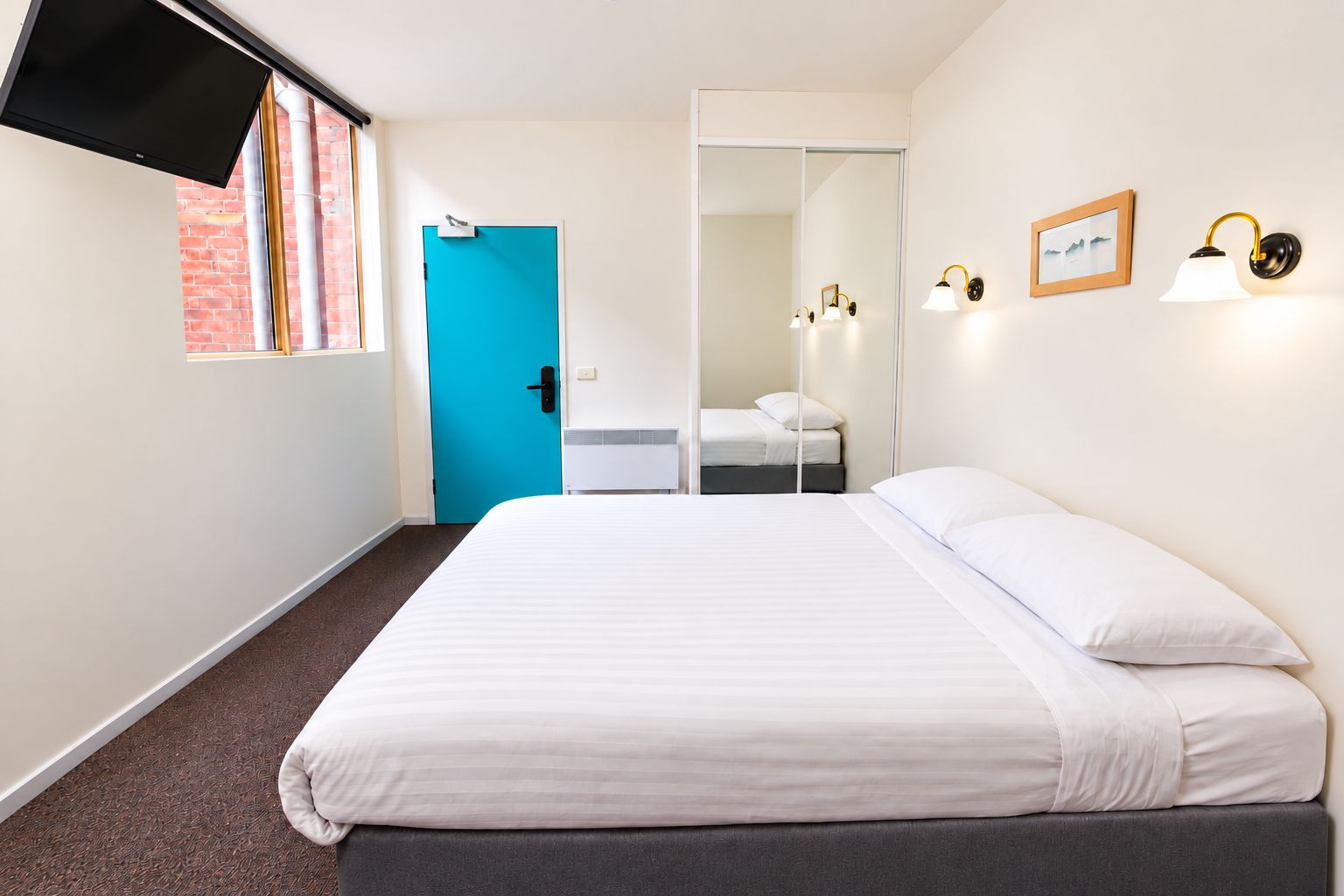 A hotel room with a bed, a TV, a blue door, a mirror, and wall-mounted lights.