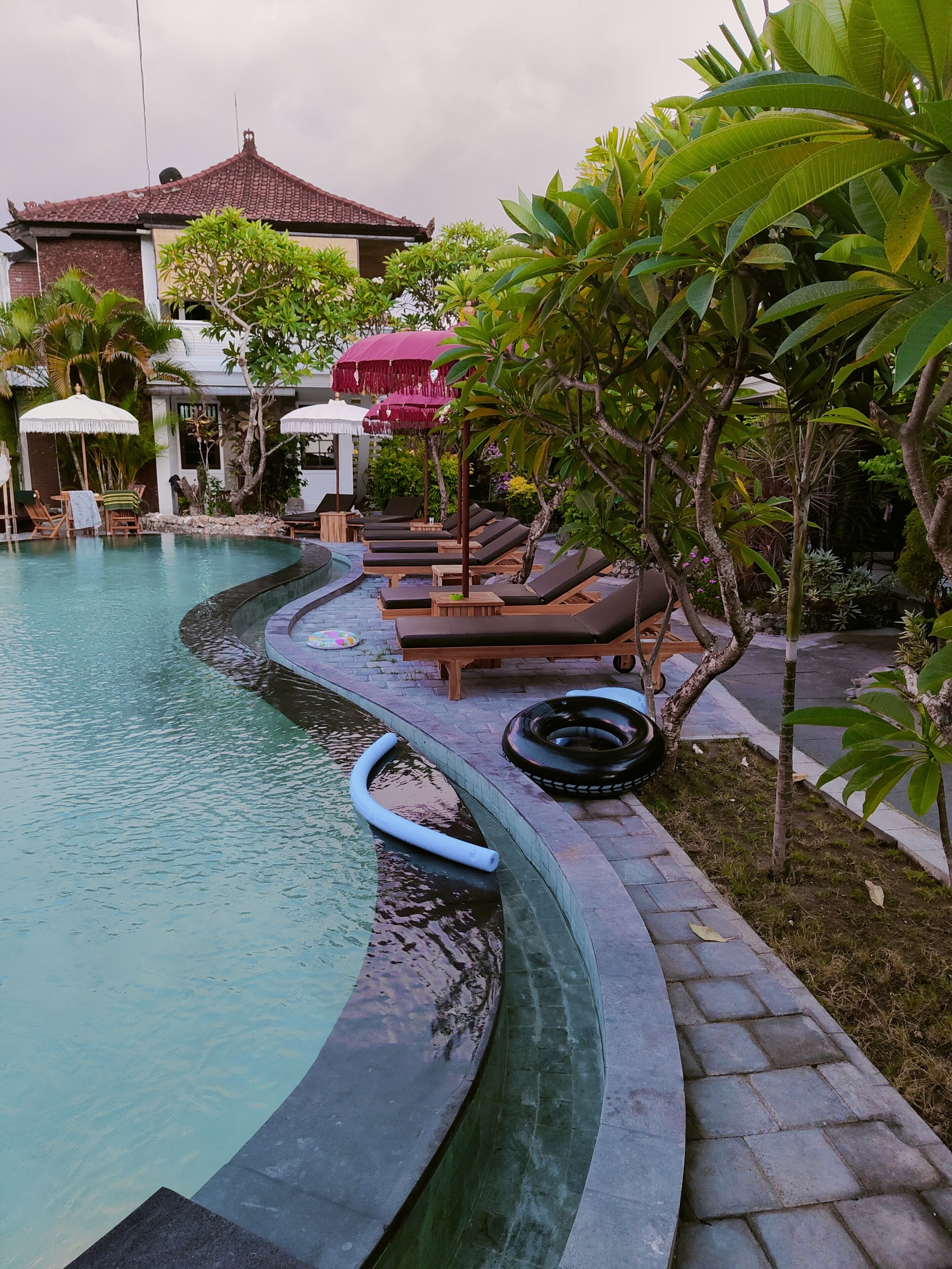 A pool area with lounge chairs and umbrellas, surrounded by tropical plants and a building in the background.