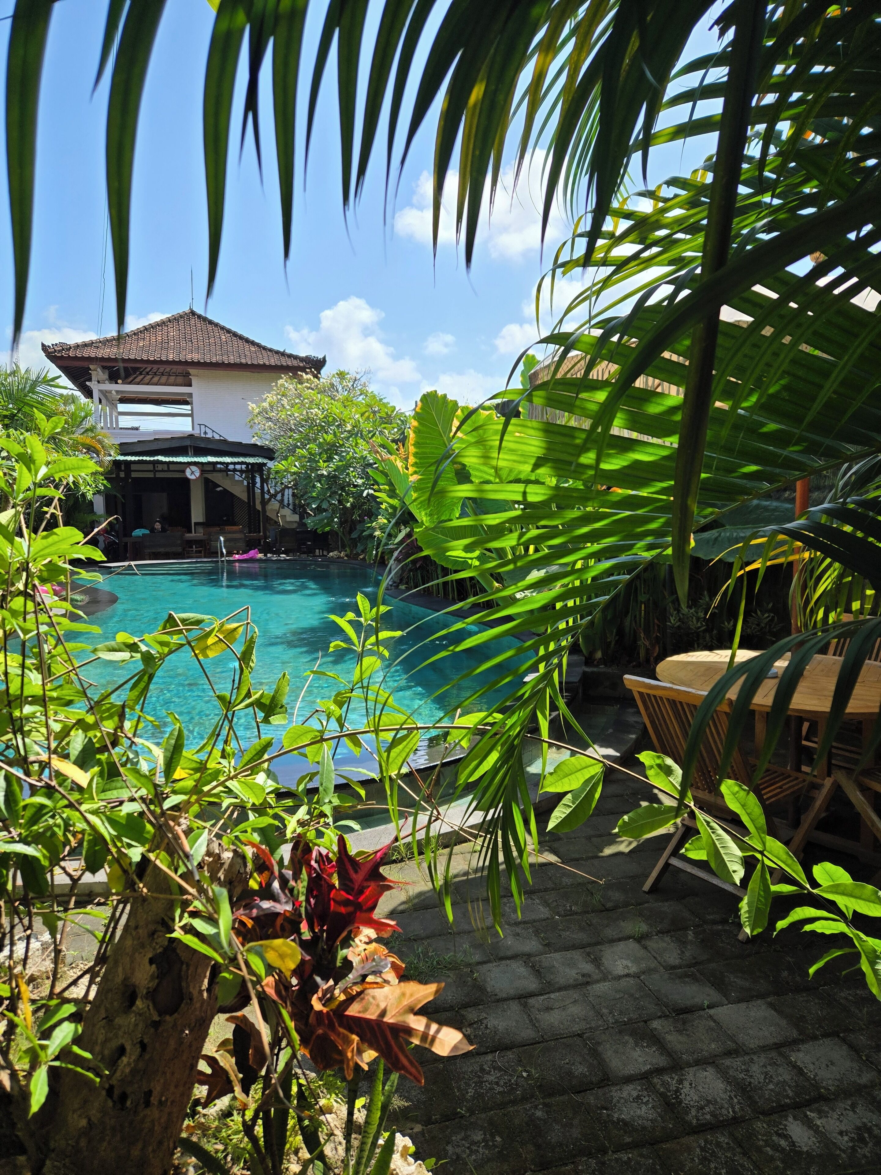 A pool area with a covered seating area, surrounded by lush greenery and a traditional building with a tiled roof.