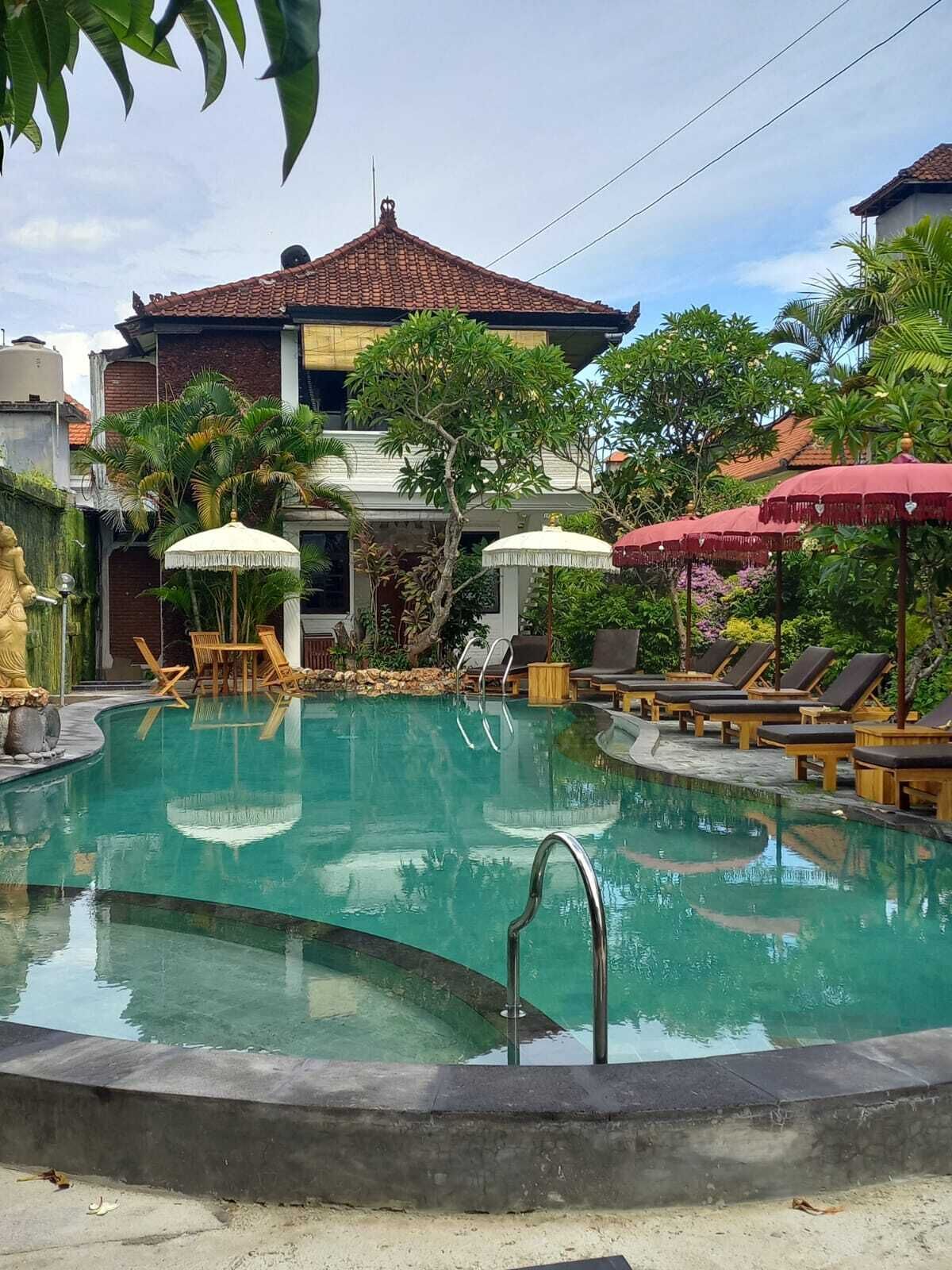 A swimming pool with lounge chairs and umbrellas, surrounded by greenery and a building with a tiled roof.