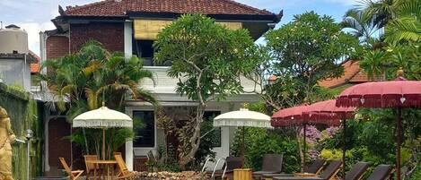 A swimming pool with lounge chairs and umbrellas, surrounded by greenery and a building with a tiled roof.