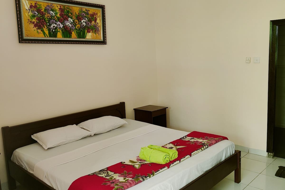 A neatly made bed with a red and white bedspread, a floral painting on the wall, and a small bedside table.
