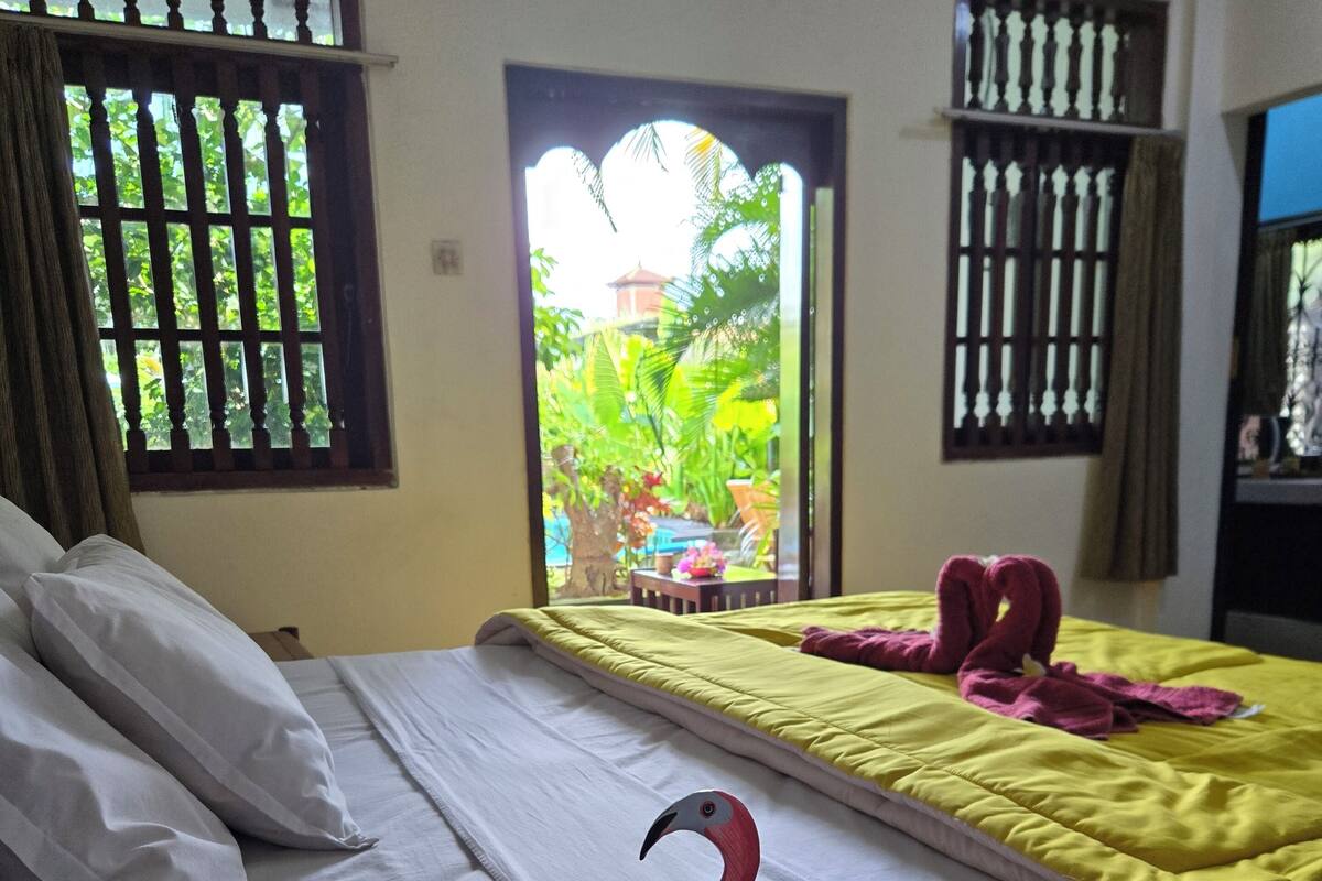 A hotel room with a bed, a window with a view of greenery, and a decorative flamingo on the bed.