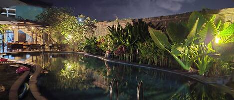 A nighttime pool area with a clear pool, surrounding greenery, and a building with outdoor seating.