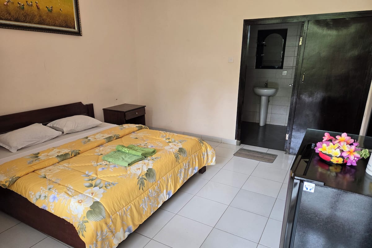 A neatly made bed with floral bedding, a bedside table, a small cabinet, and a bathroom visible through an open door.