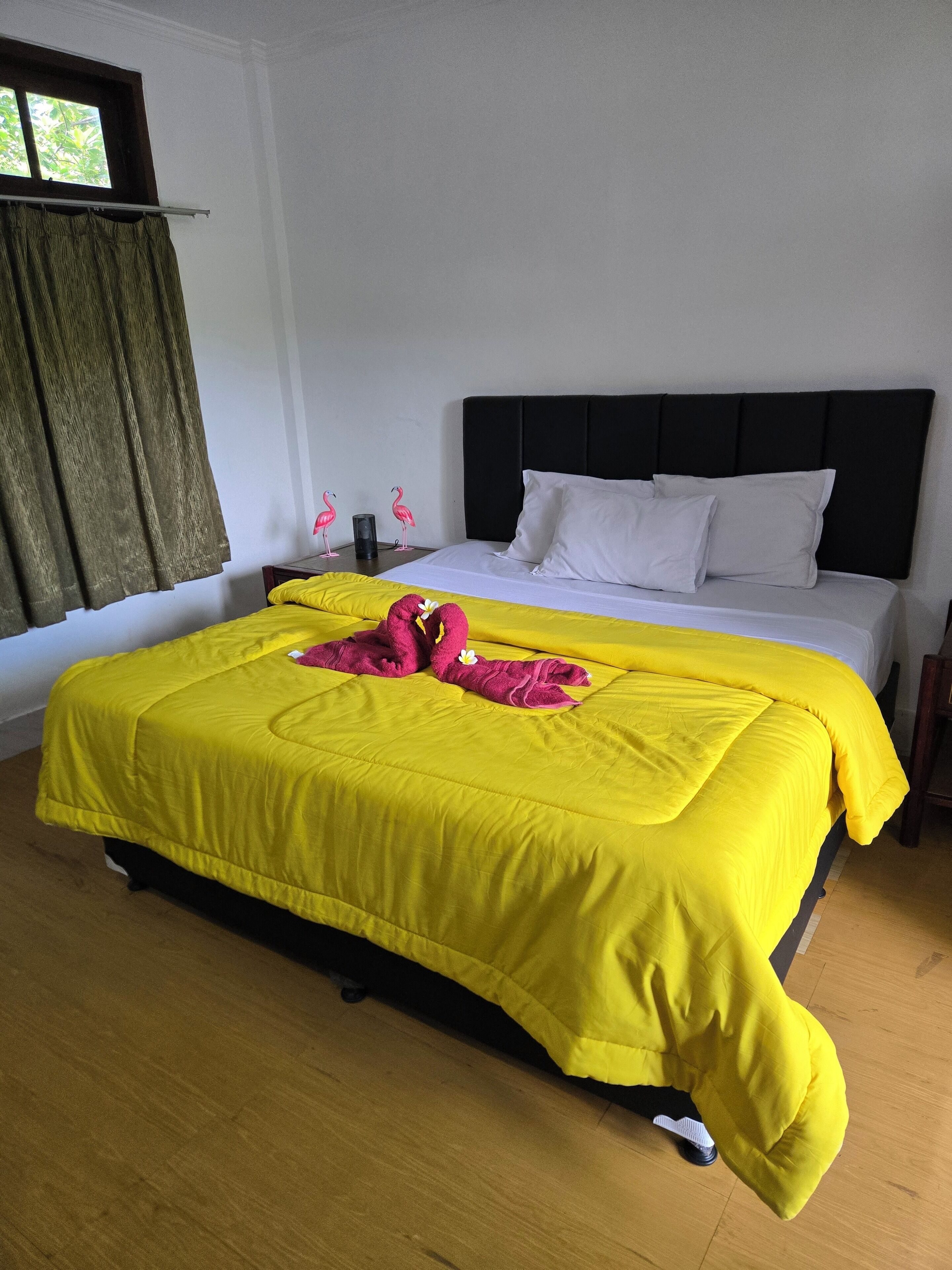 A hotel room with a bed, a window with curtains, and a nightstand with two flamingo figurines.