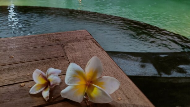 Une table en bois, ornée de deux fleurs, avec vue sur une piscine et un bâtiment doté de parasols et de chaises longues.