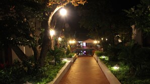 A nighttime pathway flanked by trees and illuminated by street lamps, leading to a building with a covered entrance.